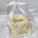 Small decorative cake with 'forever' text in a clear box with a white ribbon on a white background