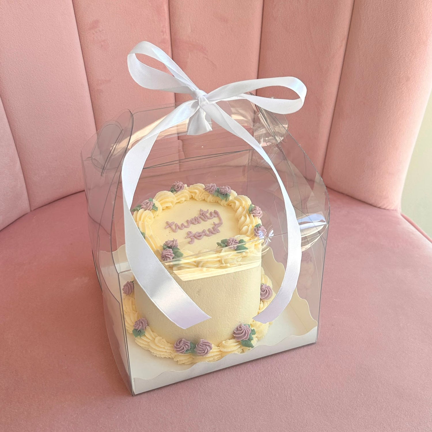 A small round birthday cake with yellow frosting and purple floral decorations, packaged in a clear square plastic box tied with a white ribbon, set against a pink pleated fabric background.